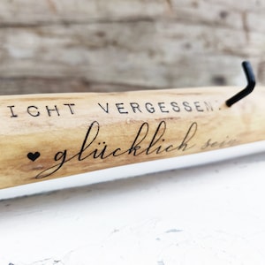 Könnte beinhalten: Ein Holzschild mit dem Text "NICHT VERGESSEN: glücklich sein" und einem kleinen Herzsymbol. Das Schild hat einen schwarzen Metallhaken.