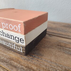 Puede incluir: Un letrero de bloques de madera con el texto "all is proof that change is beautiful" en letras blancas. Los bloques est&aacute;n apilados en tres capas, con la capa superior pintada de color melocot&oacute;n claro, la capa intermedia de color blanco y la capa inferior de color marr&oacute;n oscuro.