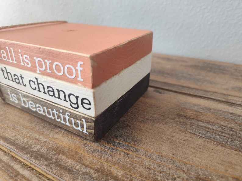Puede incluir: Un letrero de bloques de madera con el texto "all is proof that change is beautiful" en letras blancas. Los bloques est&aacute;n apilados en tres capas, con la capa superior pintada de color melocot&oacute;n claro, la capa intermedia de color blanco y la capa inferior de color marr&oacute;n oscuro.