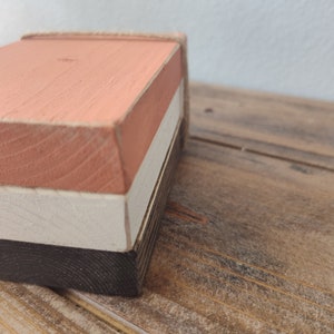Puede incluir: Una caja de madera con tres capas pintadas en tonos rosa, blanco y negro. La caja est&aacute; hecha de madera natural y tiene un aspecto r&uacute;stico.