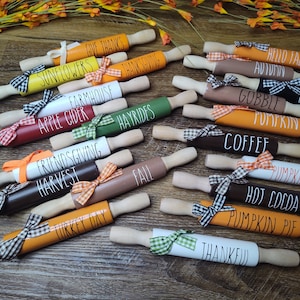 May include: A collection of 15 wooden rolling pins with various fall-themed designs and text, including "Sunflowers", "Farmhouse", "Apple Cider", "Gobble Pumpkin", "Gather", "Hayrides", "Friendsgiving Harvest", "Coffee", "Fall", "Pumpkin Spice", "Hot Cocoa", "Pumpkin Pie", and "Thankful". Each rolling pin is decorated with a plaid ribbon.