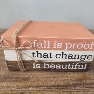 Puede incluir: Una pila de tres bloques de madera con una cita sobre el oto&ntilde;o: "fall is proof that change is beautiful". Los bloques est&aacute;n pintados en tonos de marr&oacute;n, blanco y melocot&oacute;n, y est&aacute;n atados con un cordel.