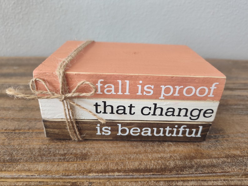 Puede incluir: Una pila de tres bloques de madera con una cita sobre el oto&ntilde;o: "fall is proof that change is beautiful". Los bloques est&aacute;n pintados en tonos de marr&oacute;n, blanco y melocot&oacute;n, y est&aacute;n atados con un cordel.