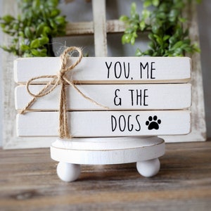 May include: White wooden sign with the text "You, Me & The Dogs" with a black paw print. The sign is stacked on a white pedestal with three legs.