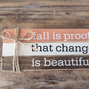 Puede incluir: Tres bloques de madera apilados uno encima del otro con una cuerda atada alrededor. El bloque superior es de color melocot&oacute;n y dice "fall is proof". El bloque del medio es blanco y dice "that change". El bloque inferior es marr&oacute;n y dice "is beautiful".