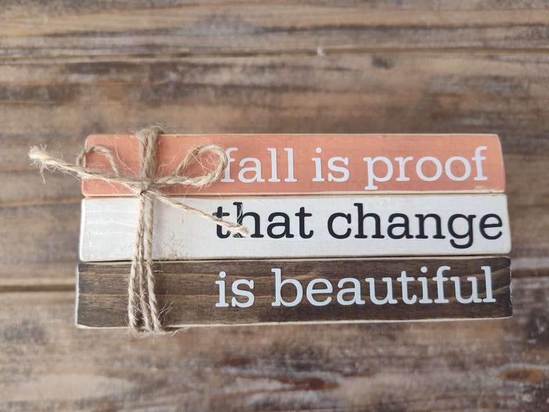 Puede incluir: Tres bloques de madera apilados uno encima del otro con una cuerda atada alrededor. El bloque superior es de color melocot&oacute;n y dice "fall is proof". El bloque del medio es blanco y dice "that change". El bloque inferior es marr&oacute;n y dice "is beautiful".