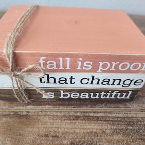 Puede incluir: Una pila de tres bloques de madera con una cita sobre el oto&ntilde;o: "fall is proof that change is beautiful". Los bloques est&aacute;n pintados en tonos de naranja, blanco y marr&oacute;n, y est&aacute;n atados con un cordel.