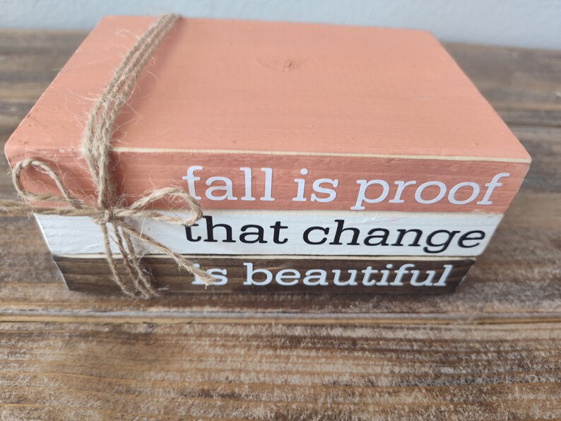 Puede incluir: Una pila de tres bloques de madera con una cita sobre el oto&ntilde;o: "fall is proof that change is beautiful". Los bloques est&aacute;n pintados en tonos de naranja, blanco y marr&oacute;n, y est&aacute;n atados con un cordel.