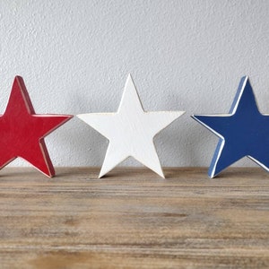 May include: Three wooden stars painted in red, white, and blue. The stars are arranged in a row on a wooden surface.
