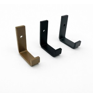 Op de afbeelding: Drie metalen wandhaken in brons, zwart en matzwart. Elke haak heeft een haakse vormgeving met een rechthoekige basis en een montagegat. De haken worden tegen een witte achtergrond getoond.