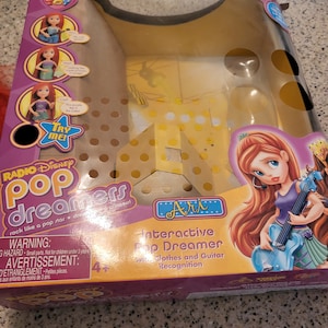 May include: A yellow and pink interactive toy with a doll and a guitar. The box says "Radio Disney Pop Dreamers" and "Interactive Pop Dreamer with Clothes and Guitar Recognition".