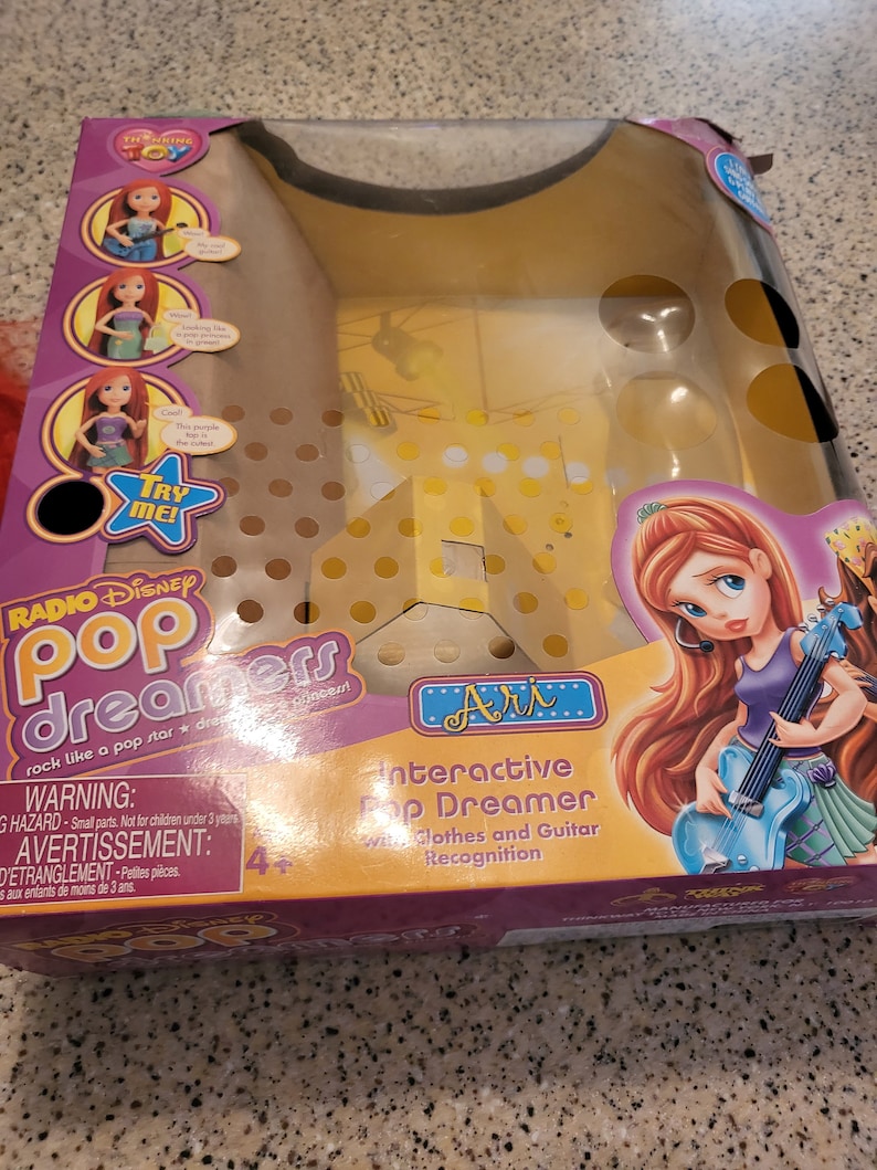 May include: A yellow and pink interactive toy with a doll and a guitar. The box says "Radio Disney Pop Dreamers" and "Interactive Pop Dreamer with Clothes and Guitar Recognition".