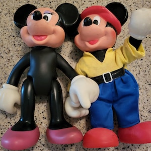 May include: A vintage pair of Mickey Mouse and Minnie Mouse dolls. Mickey is wearing a red cap, yellow shirt, blue jeans, and white gloves. Minnie is wearing a black dress, pink shoes, and white gloves.