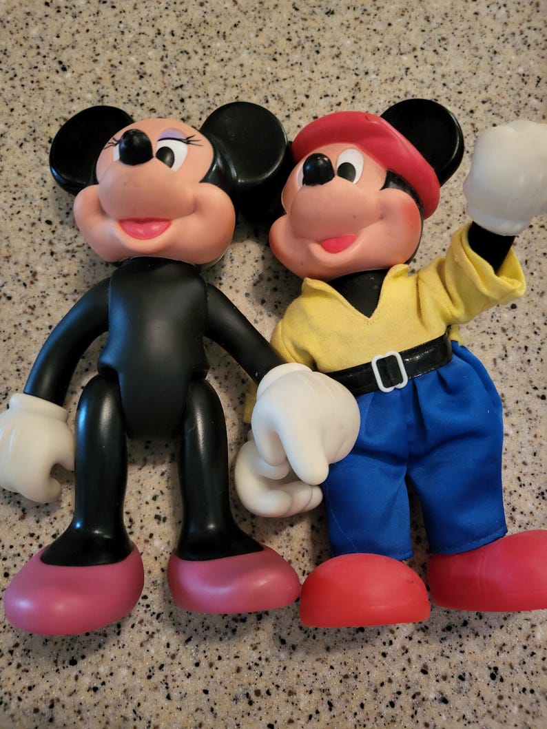 May include: A vintage pair of Mickey Mouse and Minnie Mouse dolls. Mickey is wearing a red cap, yellow shirt, blue jeans, and white gloves. Minnie is wearing a black dress, pink shoes, and white gloves.
