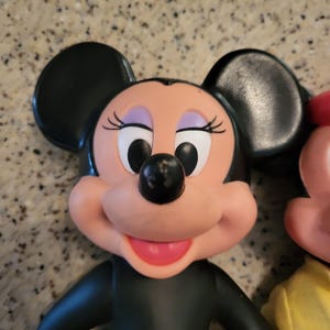May include: A vintage black and pink plastic toy figurine of Minnie Mouse. She has large black ears, a black nose, and a pink smile. Her eyes are black with white pupils.
