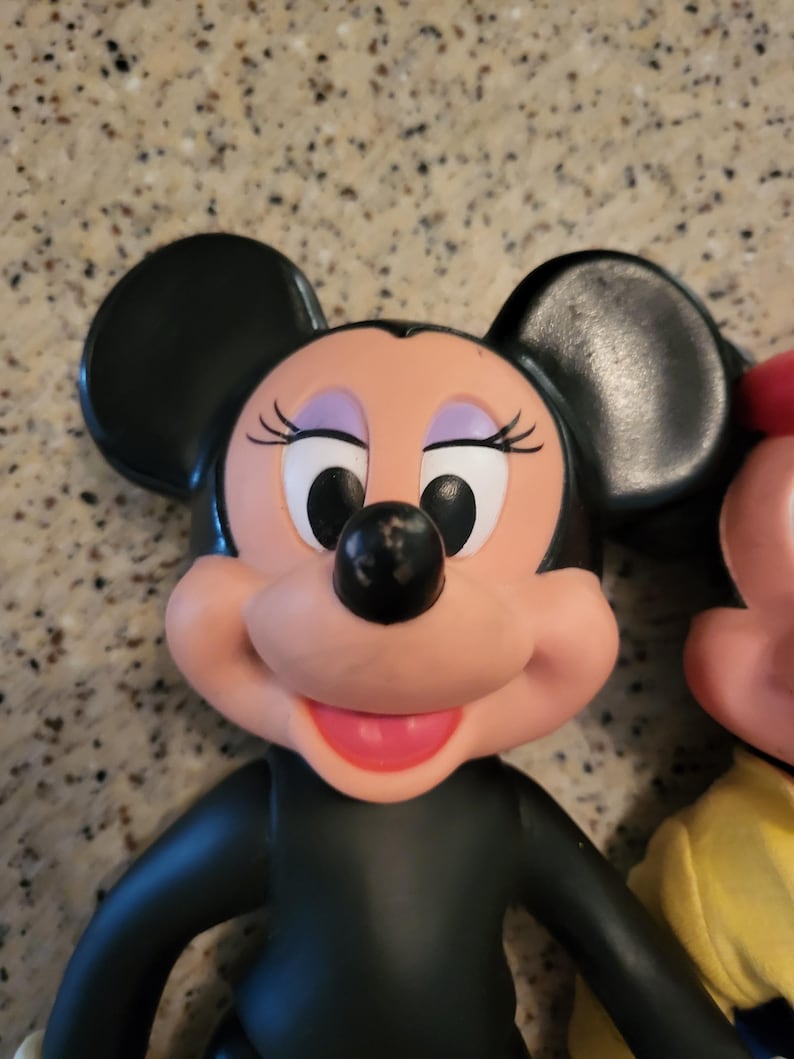 May include: A vintage black and pink plastic toy figurine of Minnie Mouse. She has large black ears, a black nose, and a pink smile. Her eyes are black with white pupils.
