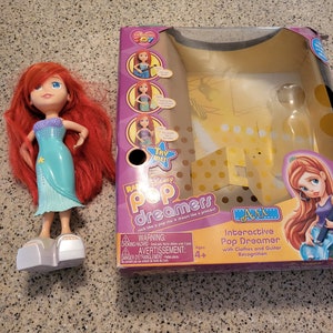 May include: A red-haired doll wearing a teal mermaid dress and silver shoes. The doll is standing on a table. The doll is next to a box with a picture of a girl with a guitar. The box says "Interactive Pop Dreamer with Clothes and Guitar Recognition".