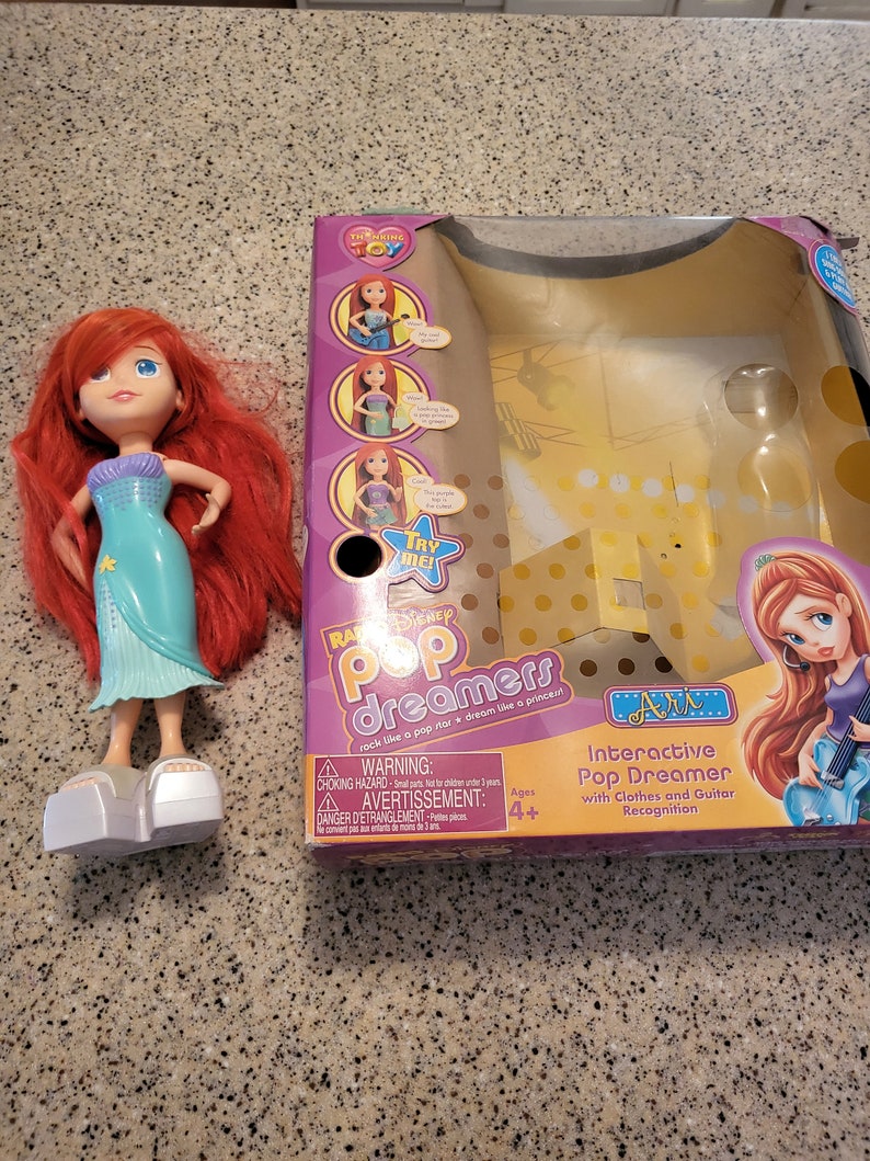 May include: A red-haired doll wearing a teal mermaid dress and silver shoes. The doll is standing on a table. The doll is next to a box with a picture of a girl with a guitar. The box says "Interactive Pop Dreamer with Clothes and Guitar Recognition".