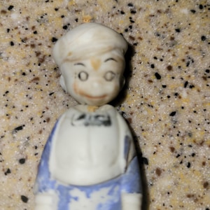 以下が含まれることがあります： 小さな、ヴィンテージの磁器製の少年の置物。置物は白い帽子とシャツ、青いオーバーオールを着ています。顔は、目、鼻、笑顔の口など、繊細な特徴で描かれています。置物はコレクターアイテムです。
