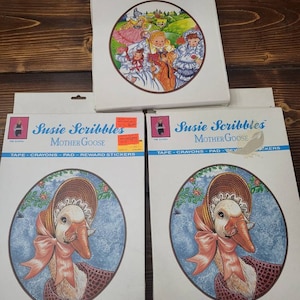 Puede incluir: Tres cajas de libros de actividades Susie Scribbles. La caja superior presenta una ilustración colorida de niños en un campo. Las dos cajas inferiores presentan una oca con un gorro y una cinta rosa. El texto en las cajas dice "Susie Scribbles Nursery Tales" y "Susie Scribbles Mother Goose".