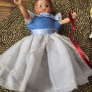 May include: A porcelain doll wearing a white dress with a blue bodice and a red ribbon. The doll has a painted face with brown hair and blue eyes.