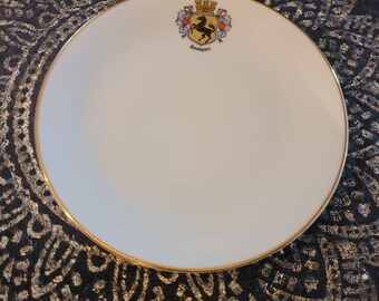 May include: A white ceramic plate with a gold rim and a black and gold crest featuring a horse and the word "Scampori". The plate is on a black and gold patterned surface.