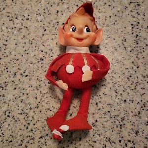 Może przedstawiać: Lalka elfika świątecznego w kolorze czerwonym i białym z dużą, okrągłą głową, spiczastym kapeluszem i długimi nogami. Elf ma uśmiechniętą twarz z dużymi oczami i szerokimi ustami.