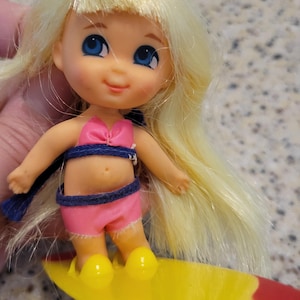 Peut inclure: Une petite poupée aux longs cheveux blonds et aux yeux bleus, portant un haut de bikini rose et un short assorti avec une bordure bleu foncé. La poupée est debout sur une planche de surf jaune et rouge, avec des chaussures moulées jaunes.