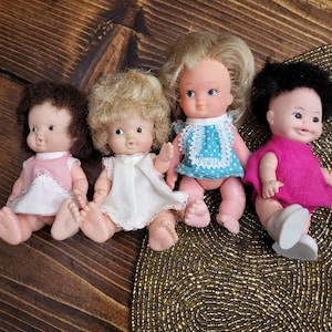 May include: Four small plastic dolls with painted faces and hair. The dolls are wearing different colored dresses and are sitting on a wooden surface.