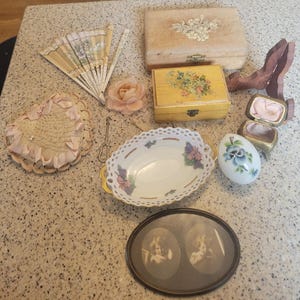 Könnte beinhalten: Eine Sammlung antiker Gegenstände, darunter ein Fächer, eine herzförmige Deckchen, eine dekorative Schale und kleine Schachteln. Der Fächer ist beige mit einem floralen Design. Die Schale ist weiß mit einem gewellten Rand und floralen Akzenten. Ein gerahmtes ovales Bild ist ebenfalls vorhanden.