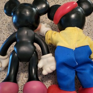 May include: Two Mickey Mouse plush toys. One is black with red shoes and the other is wearing a red hat, yellow shirt, blue pants, and red shoes.
