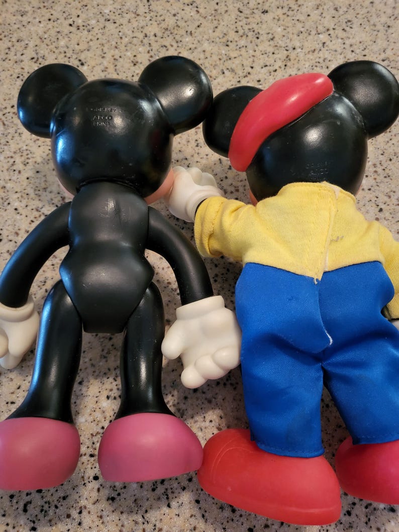 May include: Two Mickey Mouse plush toys. One is black with red shoes and the other is wearing a red hat, yellow shirt, blue pants, and red shoes.