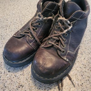 Puede incluir: Un par de botines de cuero marrón con cordones. Las botas tienen un aspecto desgastado y están atadas con cordones marrones.