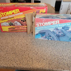 May include: Two boxes for the Stomper Action Track System toy. The box on the left is red and features a yellow toy truck on a brown bridge. The box on the right is blue and features a green toy military vehicle driving over a bridge.