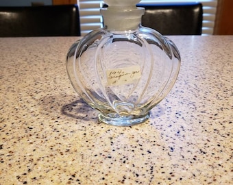 May include: A clear glass bottle with a frosted glass stopper. The bottle has a heart shape and a textured surface. A label on the bottle reads "1941 Dragon  You".