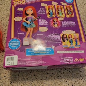 May include: A pink and purple box featuring a red-haired doll playing a blue guitar. The box is labelled "Radio Disney Pop Dreamers" and shows the doll in various outfits. It also details the doll's interactive features, including singing, playing guitar, and interacting with other dolls.
