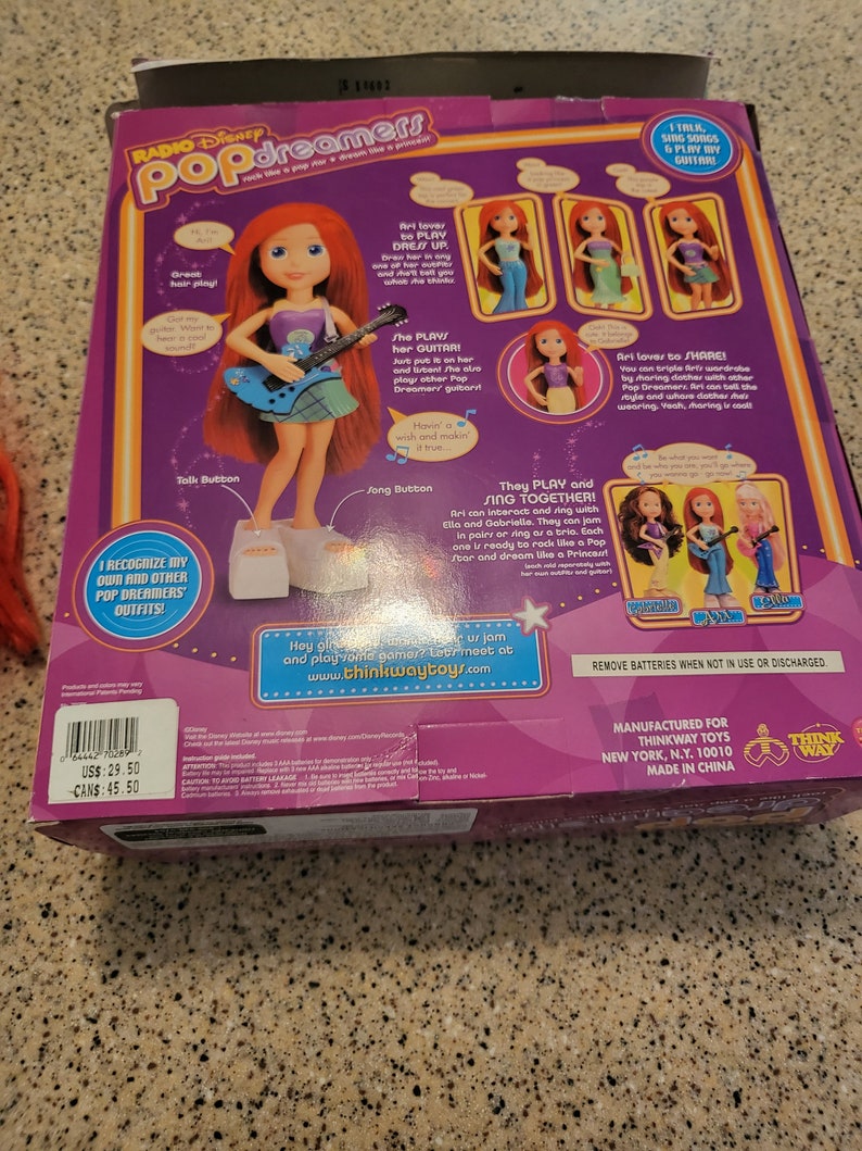 May include: A pink and purple box featuring a red-haired doll playing a blue guitar. The box is labelled "Radio Disney Pop Dreamers" and shows the doll in various outfits. It also details the doll's interactive features, including singing, playing guitar, and interacting with other dolls.