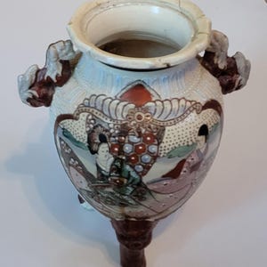 May include: A ceramic tripod vase featuring a cream-colored body with a scalloped rim and two dark brown sculpted animal-head handles. The exterior is decorated with hand-painted figures in traditional attire and a central red and blue honeycomb pattern.