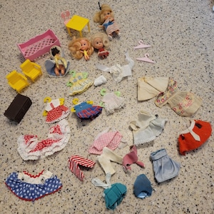 Peut inclure: Un ensemble d'accessoires de poupée miniatures comprenant trois petites poupées aux cheveux blonds et bruns, une table jaune, deux chaises jaunes, un lit rose, un étui noir, deux cintres roses et divers vêtements de poupée comme des robes et des jupes.