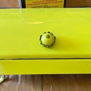 May include: A yellow metal serving tray with a decorative knob in the center. The tray has a rounded top and a flat bottom. The knob is a green and yellow flower shape.