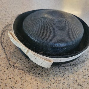 May include: A black straw hat with a cream-colored ribbon and a black netting veil. The hat has a circular crown and a wide brim. The cream-colored ribbon is tied in a bow on the side of the hat. A vintage accessory.