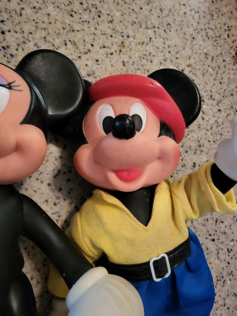 May include: A vintage Mickey Mouse and Minnie Mouse plush toy set. Mickey is wearing a red hat, yellow shirt, blue pants, and a black belt. Minnie is wearing a black dress and has a red bow on her head.