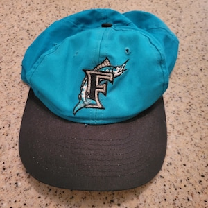 May include: A teal blue baseball cap with a black brim. The cap features an embroidered logo of a marlin fish with the letter "F" on it.