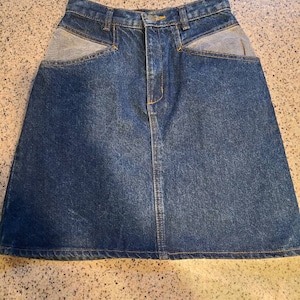 Può includere: Una gonna di jeans blu con due grandi tasche sul davanti. Le tasche sono foderate con denim blu chiaro.