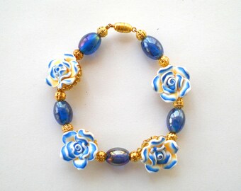 Puede incluir: Un brazalete floral azul y blanco con detalles dorados. El brazalete está hecho de cuentas de vidrio y presenta cuatro rosas blancas con detalles azules.