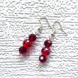 May include: A pair of dangle earrings with three red glass beads on each earring. The earrings have silver hooks.