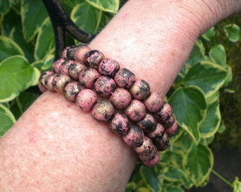 Puede incluir: Un brazalete hecho de cuentas de madera rosa y negra con detalles dorados. Las cuentas están apiladas en tres filas y tienen un acabado moteado.
