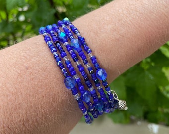 Puede incluir: Un brazalete de múltiples hilos hecho de cuentas azules y plateadas. El brazalete tiene un dije de concha.