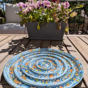 May include: A round ceramic plate featuring a series of concentric raised rings. The surface is finished in a light blue glaze with a speckled pattern of vibrant orange and dark blue spots distributed across the entire textured surface.