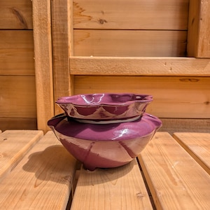 Puede incluir: Juego de cerámica de dos piezas en un tono morado intenso. El cuenco más grande tiene forma redondeada con tres patas pequeñas, mientras que el cuenco más pequeño de arriba tiene un borde festoneado. El juego se exhibe sobre una superficie de madera.